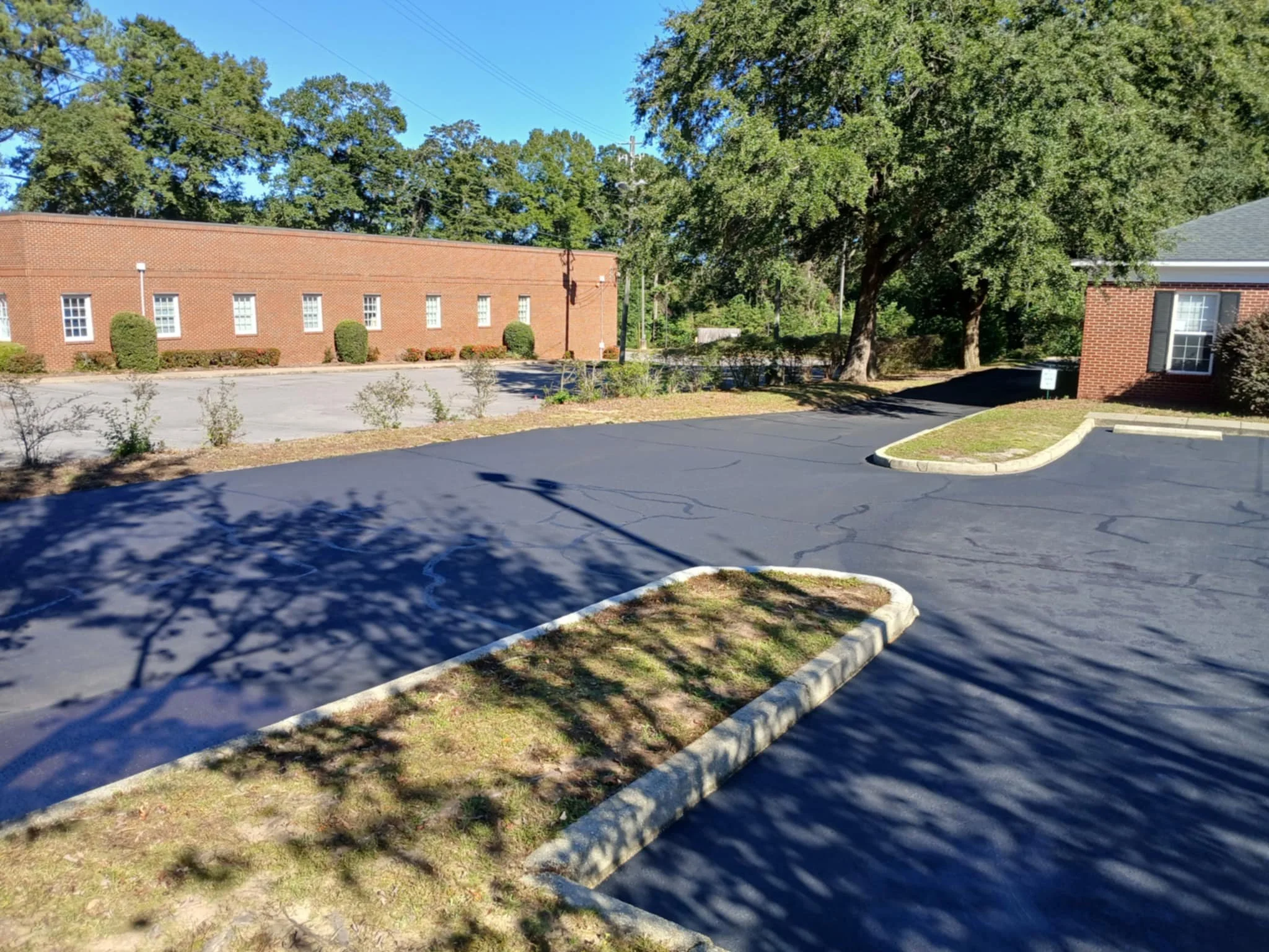 A parking lot with a small grassy island, surrounded by a brick building on the right and a larger brick building in the background. There are large trees providing shade and casting shadows on the asphalt surface.
