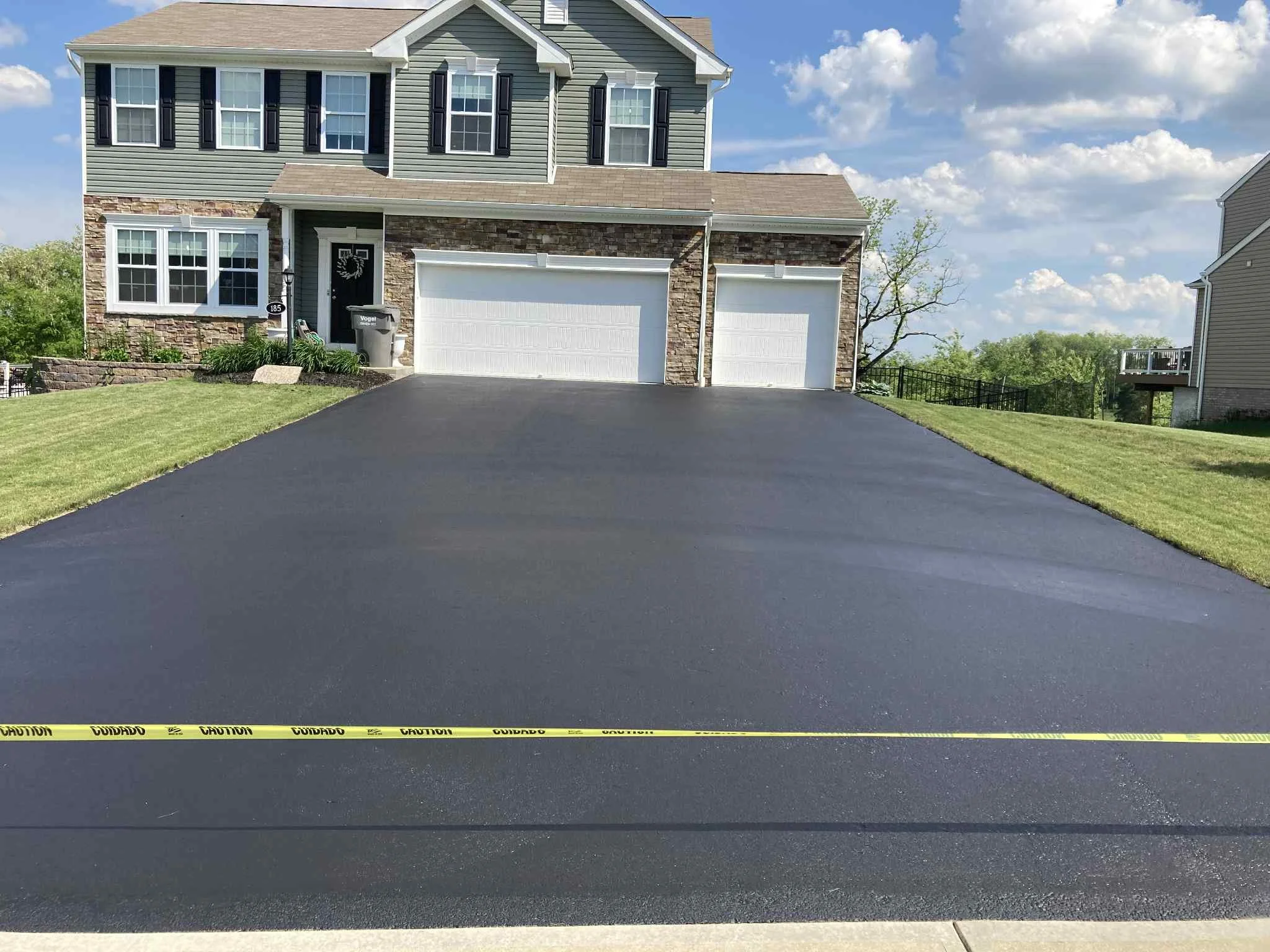 Newly paved black asphalt driveway leading to a house with a three-car garage, light green siding, stone accents, and black shutters, with green lawns on each side and caution tape across the bottom of the image.