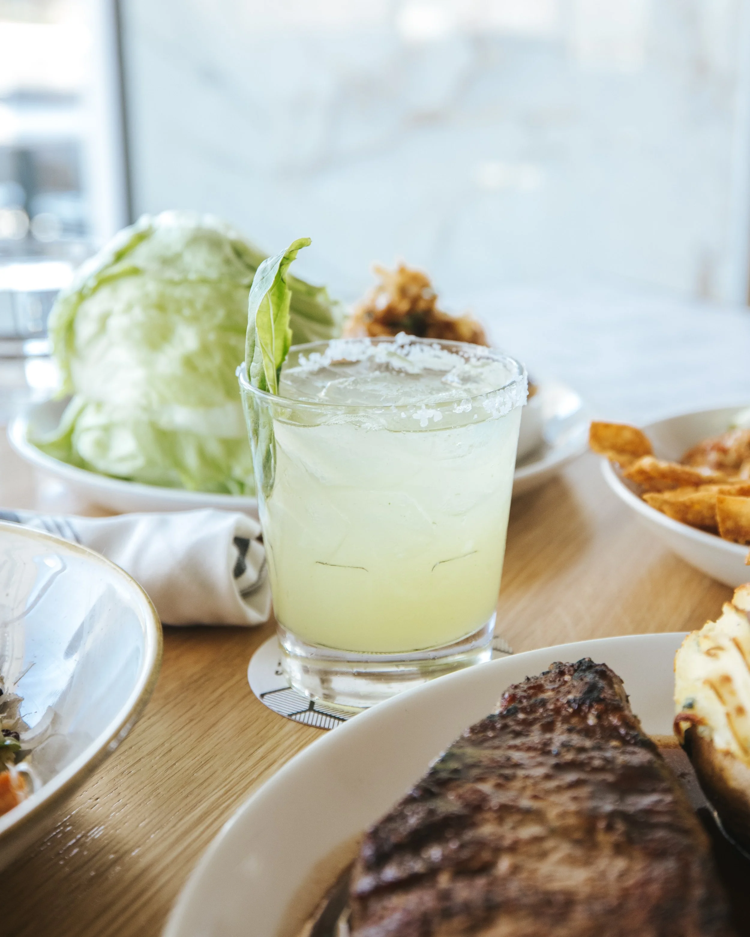 A glass of pale yellow margarita with a salted rim and lime garnish, with a bowl of shredded lettuce, fried chicken, and slices of bread or pastries in the background on a wooden table.