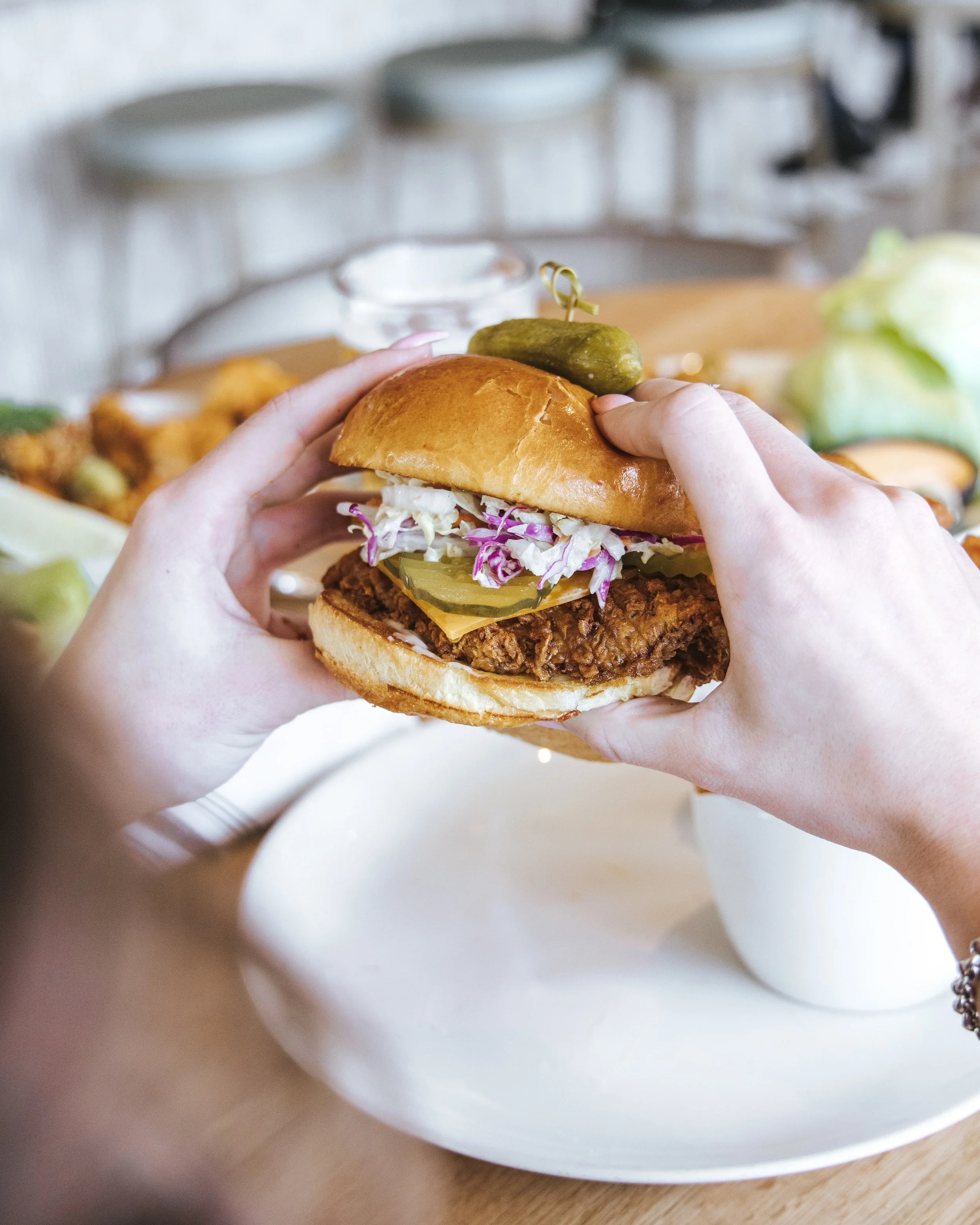 Person holding a chicken sandwich with coleslaw, pickles, cheese, and a pickle spear on top, on a white plate with a picnic table background.