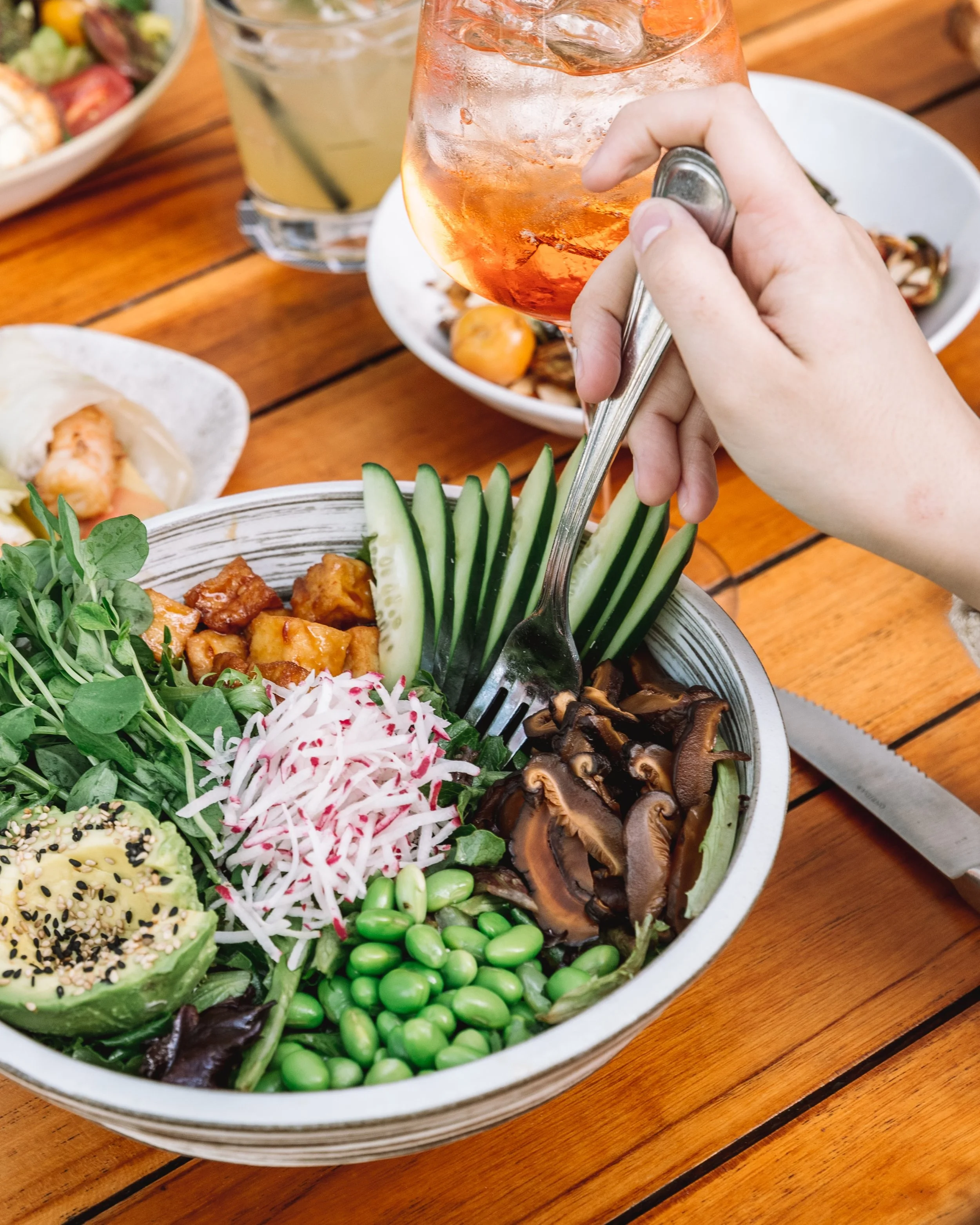 Close-up of a colorful salad with sliced avocado, radish, cucumber, edamame, mushrooms, and mixed greens in a white bowl on a wooden table, with a person's hand holding a glass of rosé wine in the background.