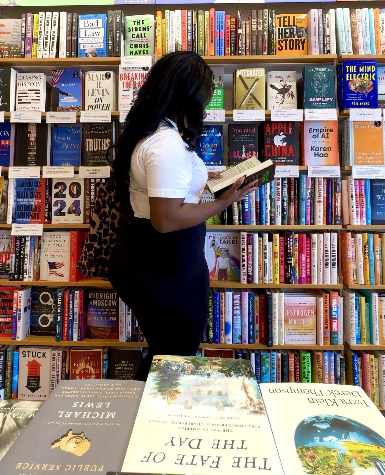 A woman with black hair wearing a white blouse and black pants, carrying a leopard print bag, is standing in front of a bookshelf in a bookstore, reading a book.
