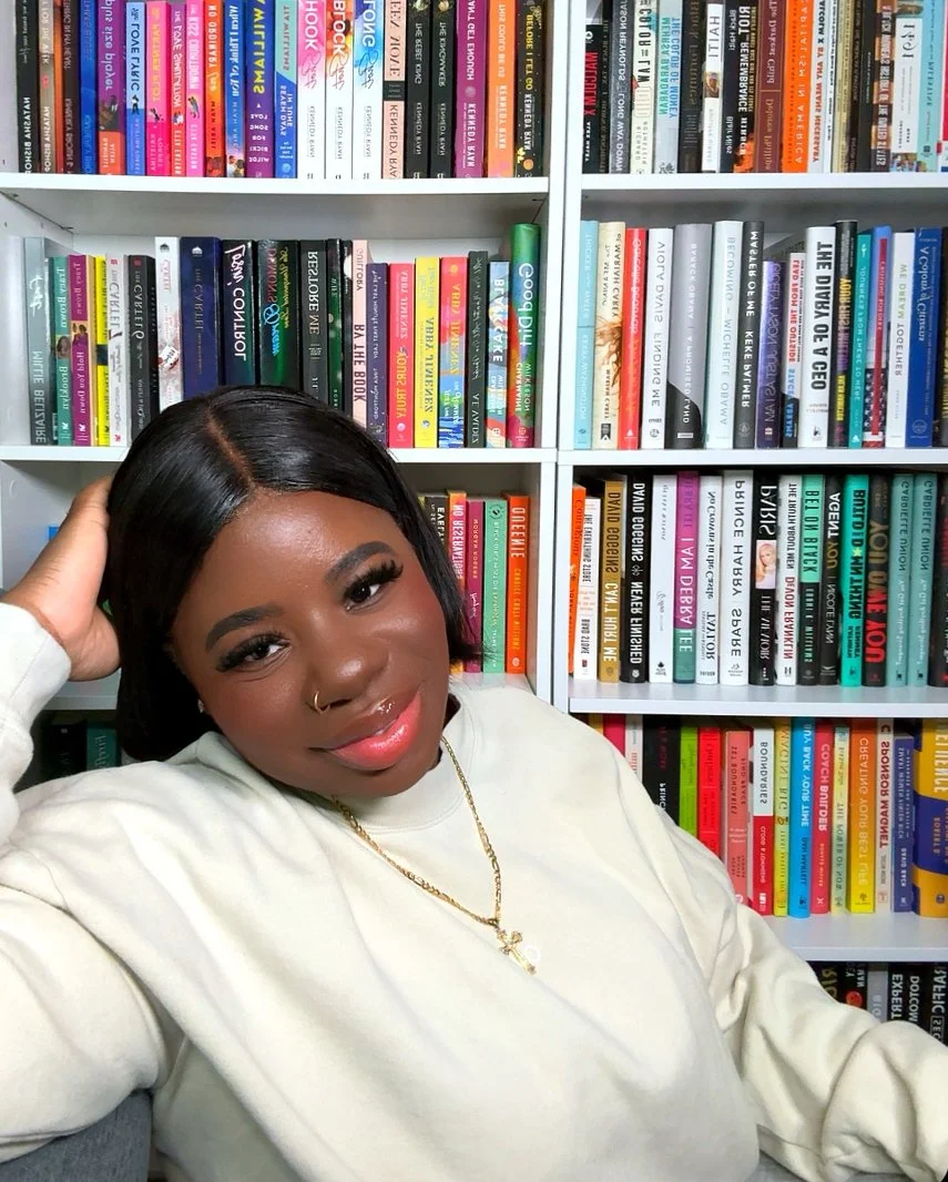 A woman with black hair, wearing a white top and gold jewelry, smiling in front of a bookshelf filled with colorful books.