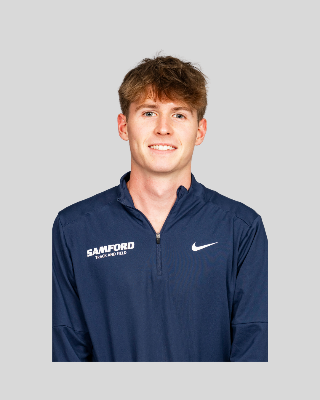 A young man with light brown hair smiling at the camera, wearing a navy blue Nike training jacket with 'Samford Track and Field' written on it, standing against a plain light gray background.
