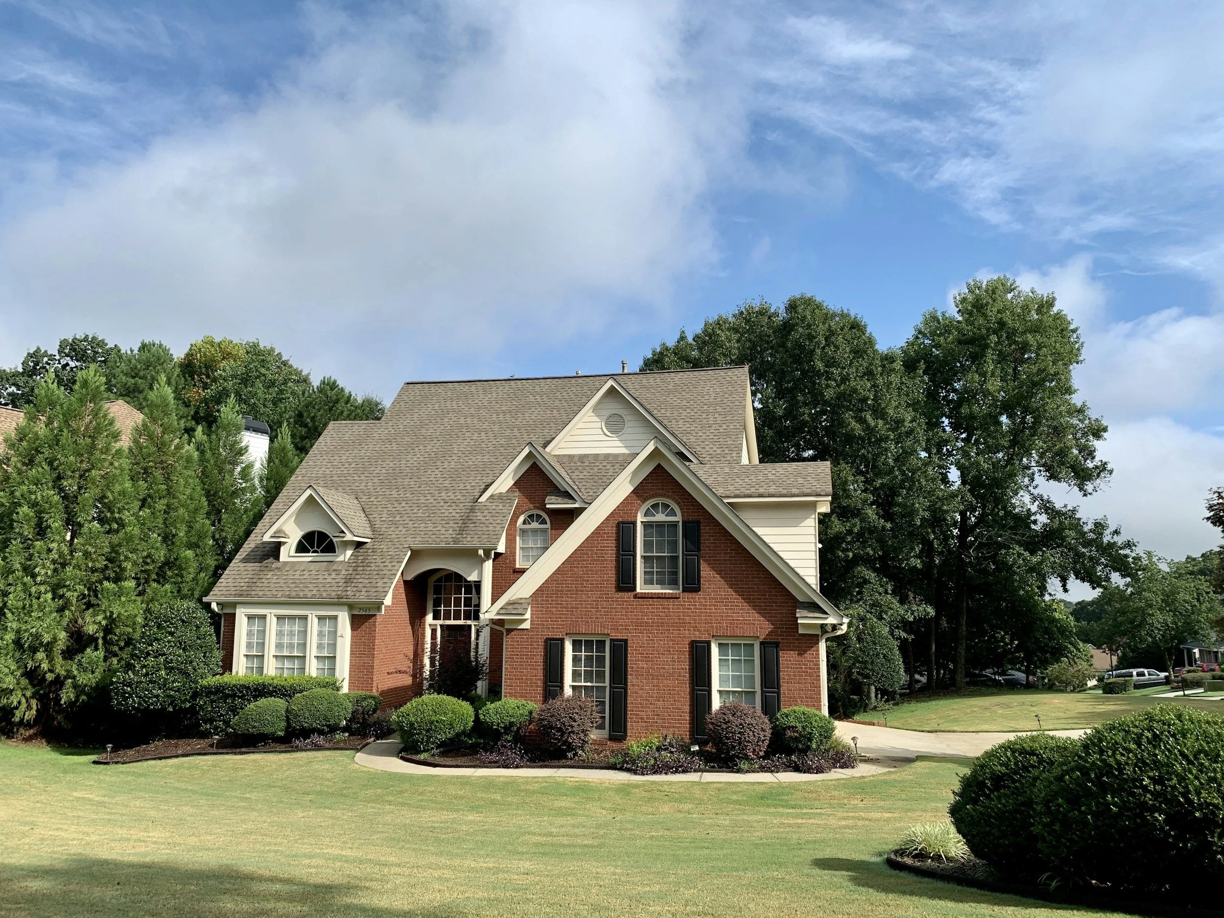 A two-story suburban house with brick and siding exterior, surrounded by well-maintained lawn and shrubs, under a partly cloudy sky.