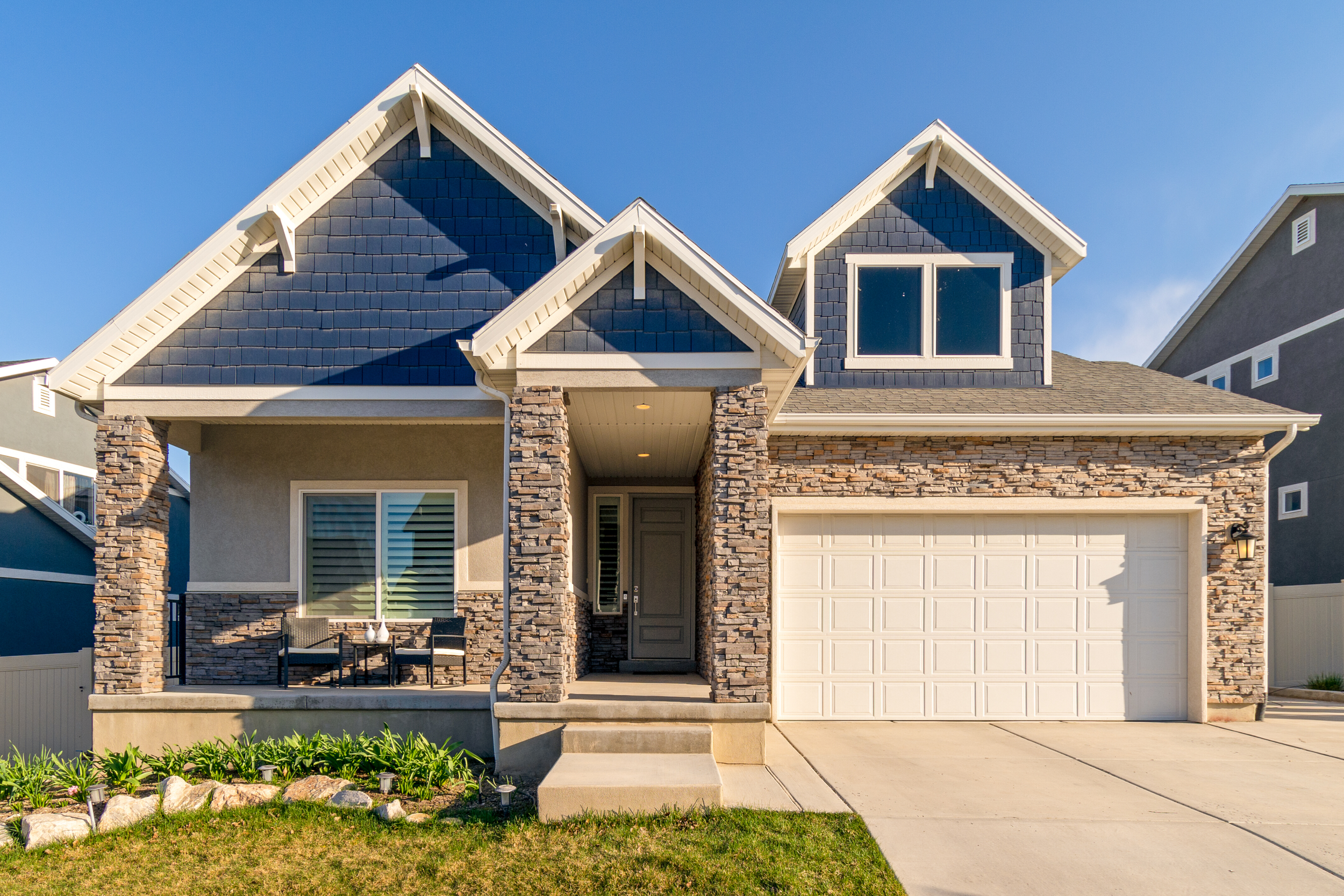 Modern house with stone accents, a porch with two chairs and a small table, a two-car garage, and a well-maintained lawn.