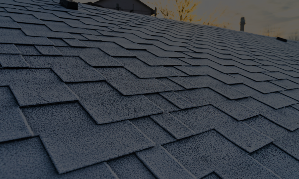 Close-up of a dark gray shingled roof with frost, sunset sky in the background.