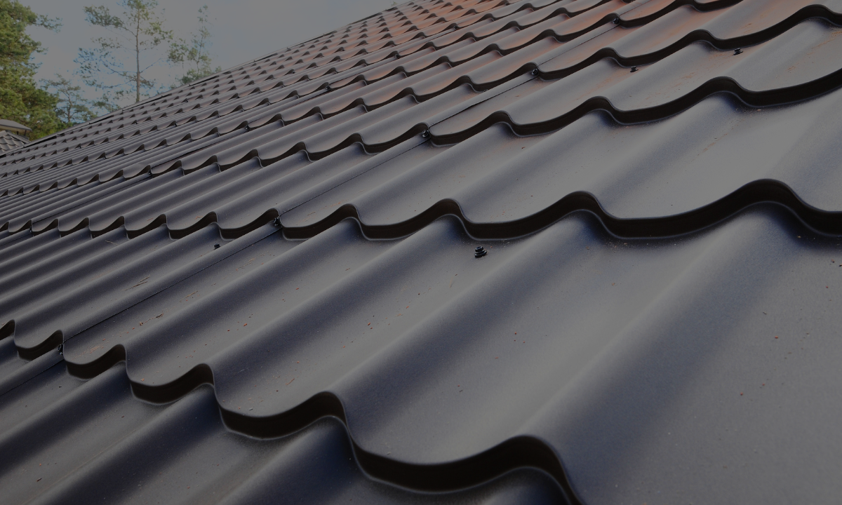 Close-up of a metal roof with wavy, ribbed panels, showing sunlight reflection and a background of clear sky and trees.