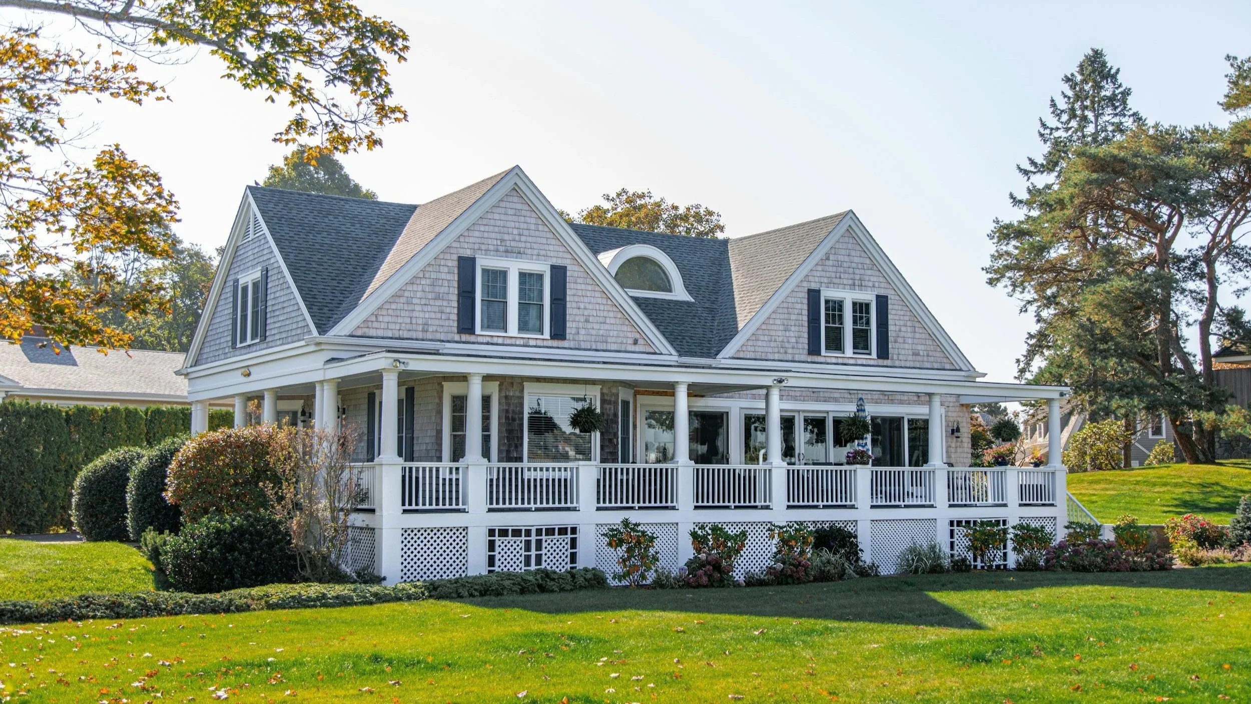A large, two-story house with a porch, white railing, and decorative shingles on the upper walls, surrounded by a well-kept yard with bushes, trees, and colorful flowers, under a clear sky.