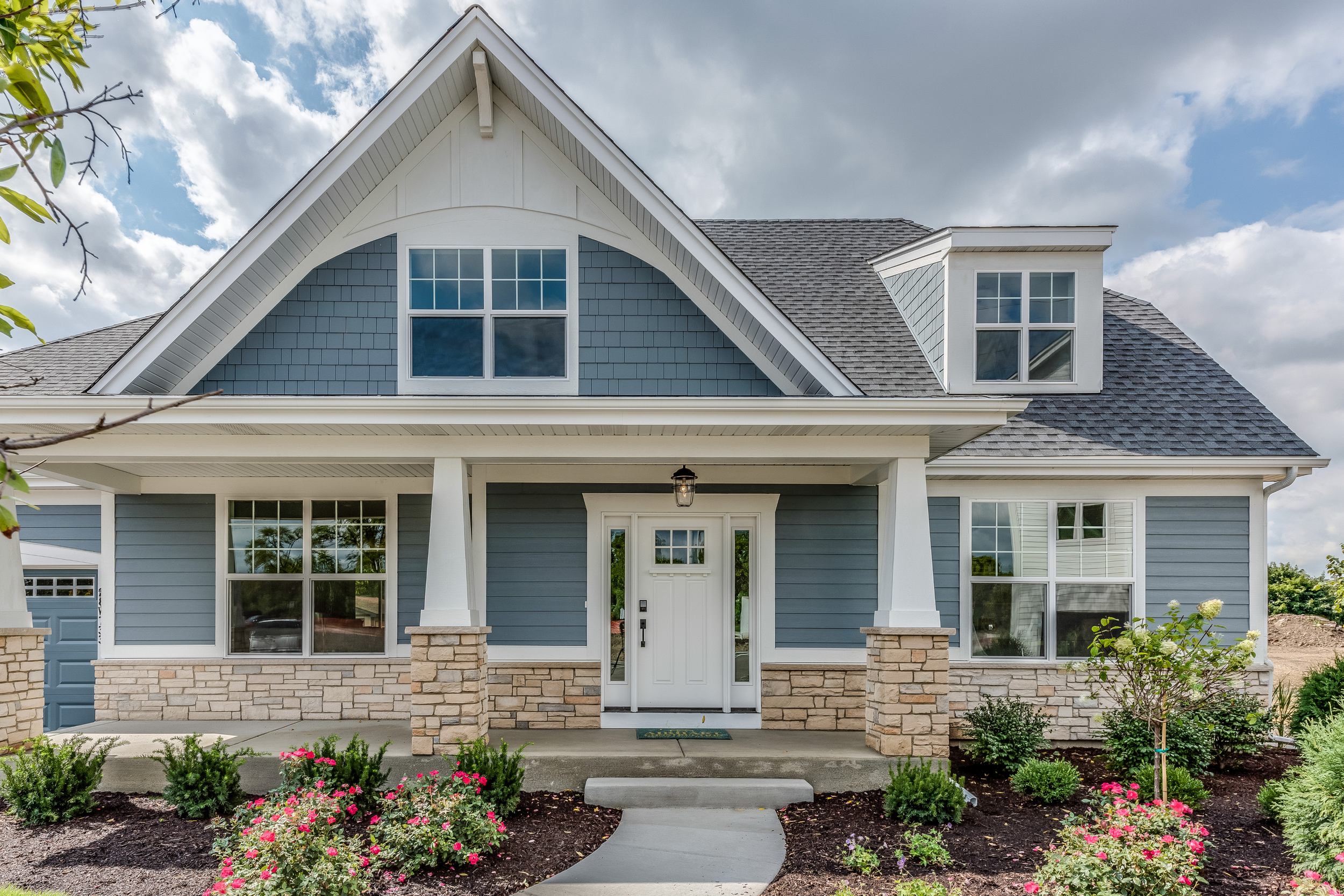 Front view of a two-story house with blue siding, white trim, and a gable roof, featuring a porch with stone pillars, a white front door, and landscaped garden with pink and white flowers.