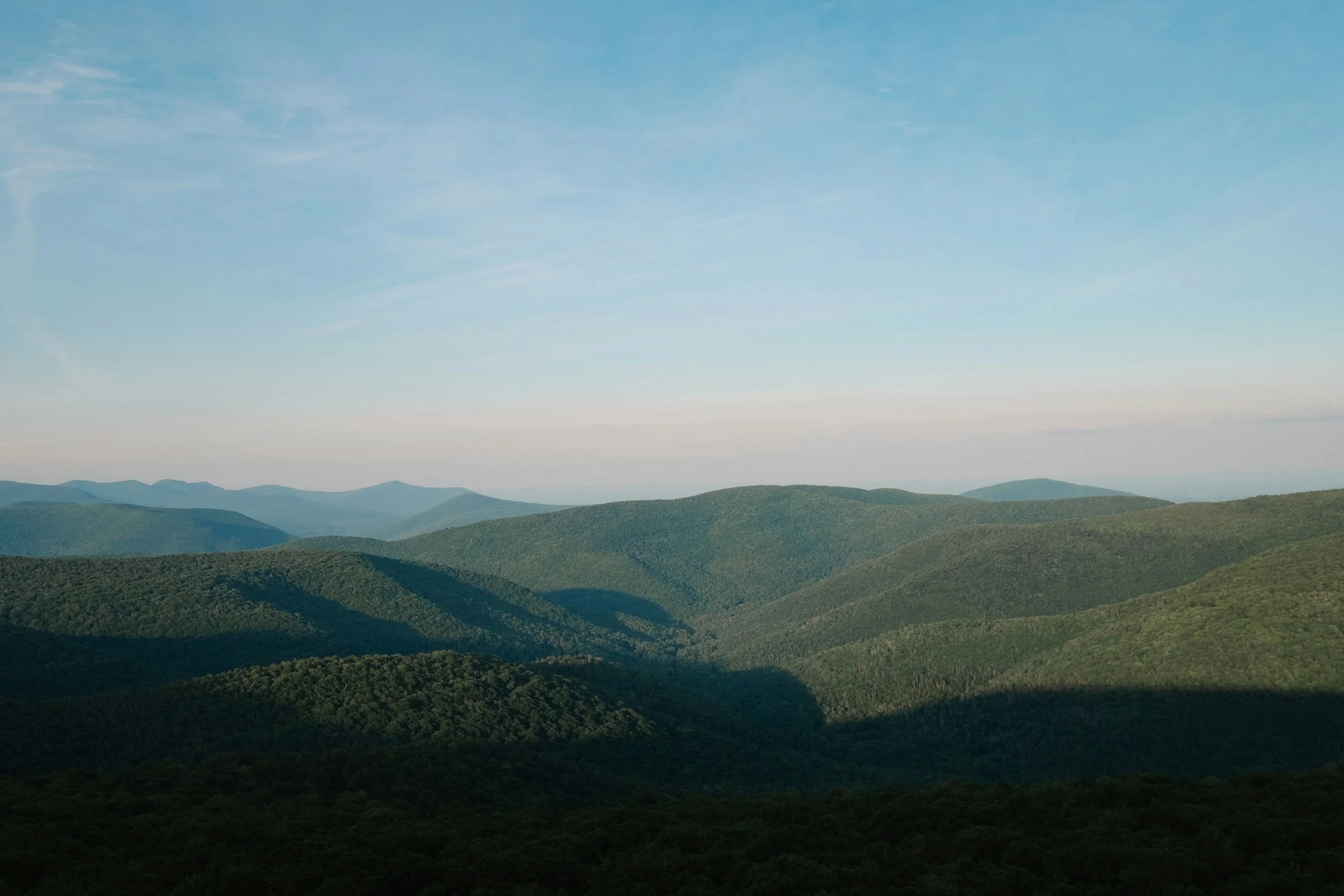 A mountainous landscape under a blue sky with light clouds, featuring green forested hills and valleys.