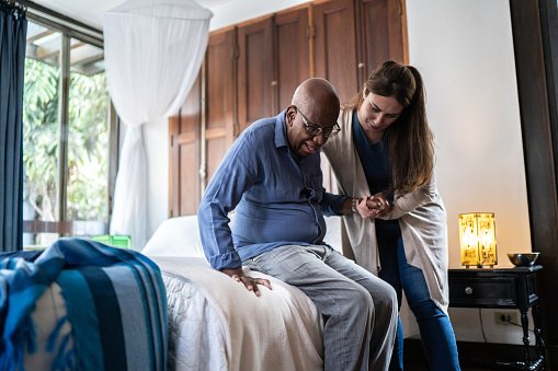 A woman assisting an elderly man with walking in a bedroom.