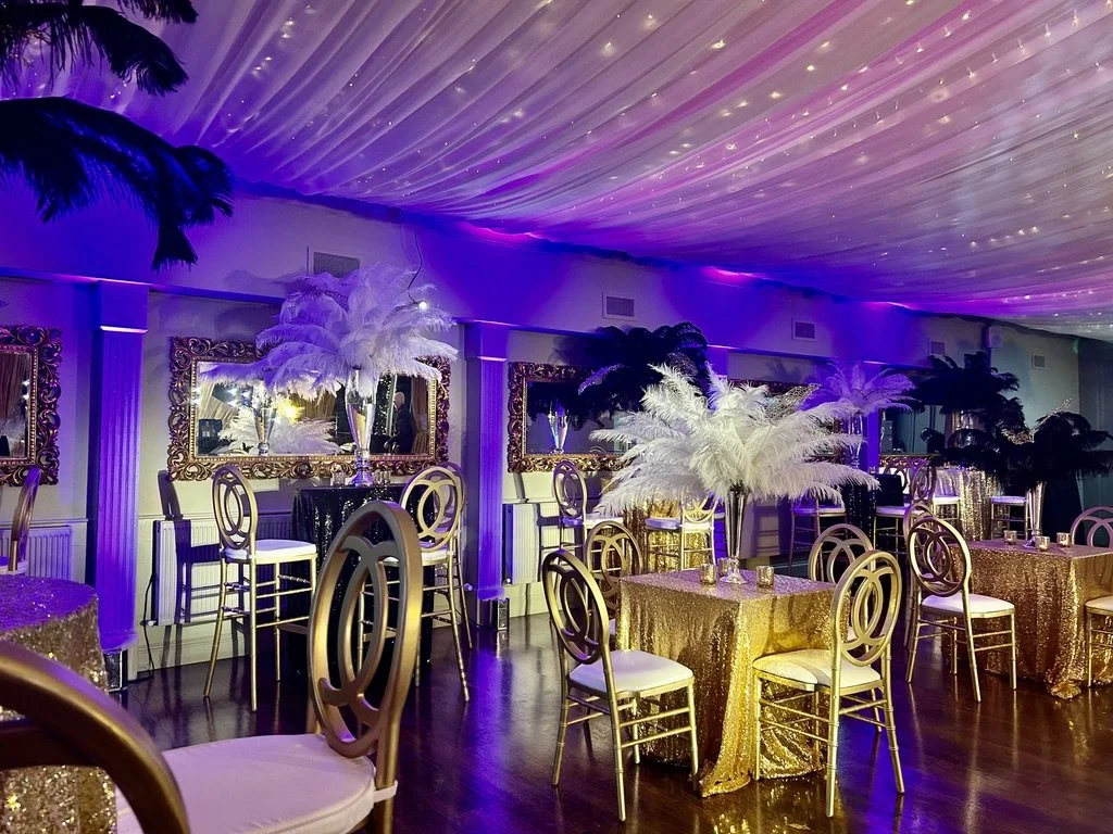 Elegant event space with tables decorated with gold sequin tablecloths, centerpieces of white and black feathers in tall vases, framed mirrors on the walls, purple lighting, and a draped ceiling.