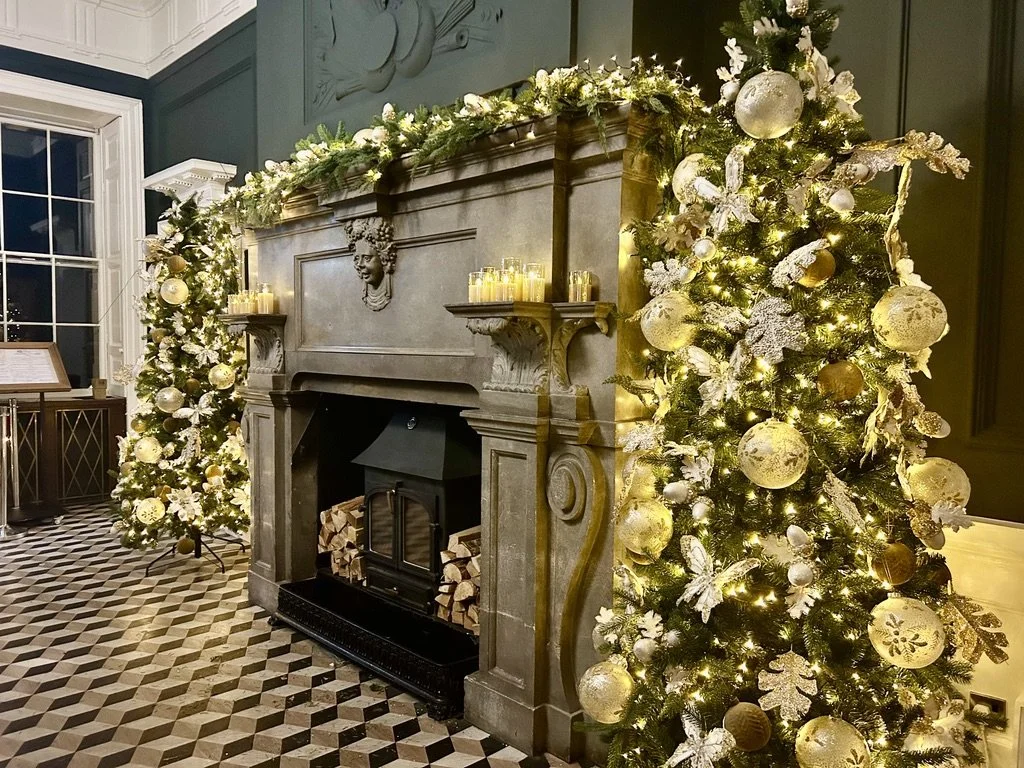Festively decorated room featuring a fireplace with candles on the mantel, two illuminated Christmas trees adorned with white and gold ornaments and lights, and a checkered tile floor.