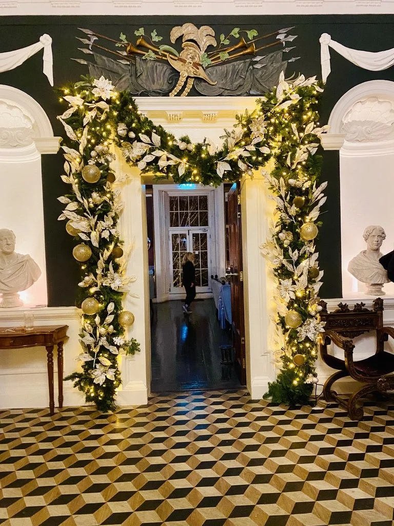 Festive holiday doorway decorated with a garland of green foliage, white leaves, and gold ornaments, illuminated with string lights, in an elegant room with bust sculptures, intricate trim, and a patterned wooden floor.