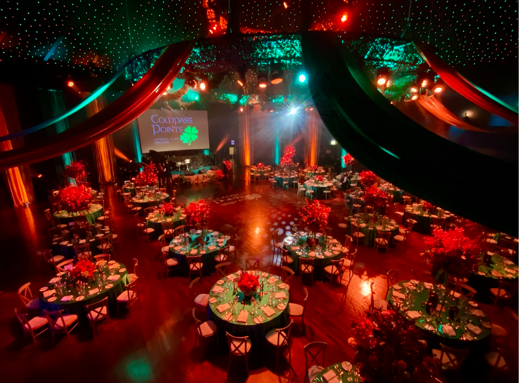 Elegant event hall decorated with round tables covered in green tablecloths, each with a bouquet of red flowers, under colorful lighting with drapery and a large screen displaying "Compass Points Ireland."