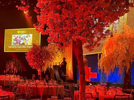 Event space with round tables and chairs, decorated with illuminated artificial trees with red, pink, and orange leaves, and a stage with speakers and a large screen.