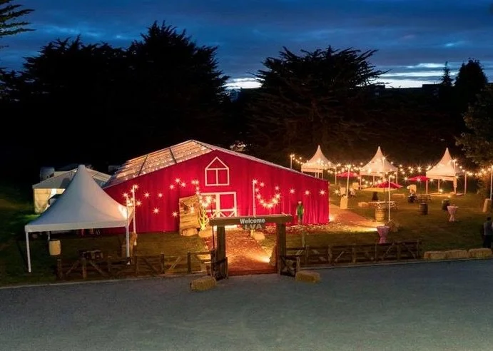 Nighttime outdoor event with a large red barn decorated with lights, surrounded by white tents with draping lights, and trees in the background.