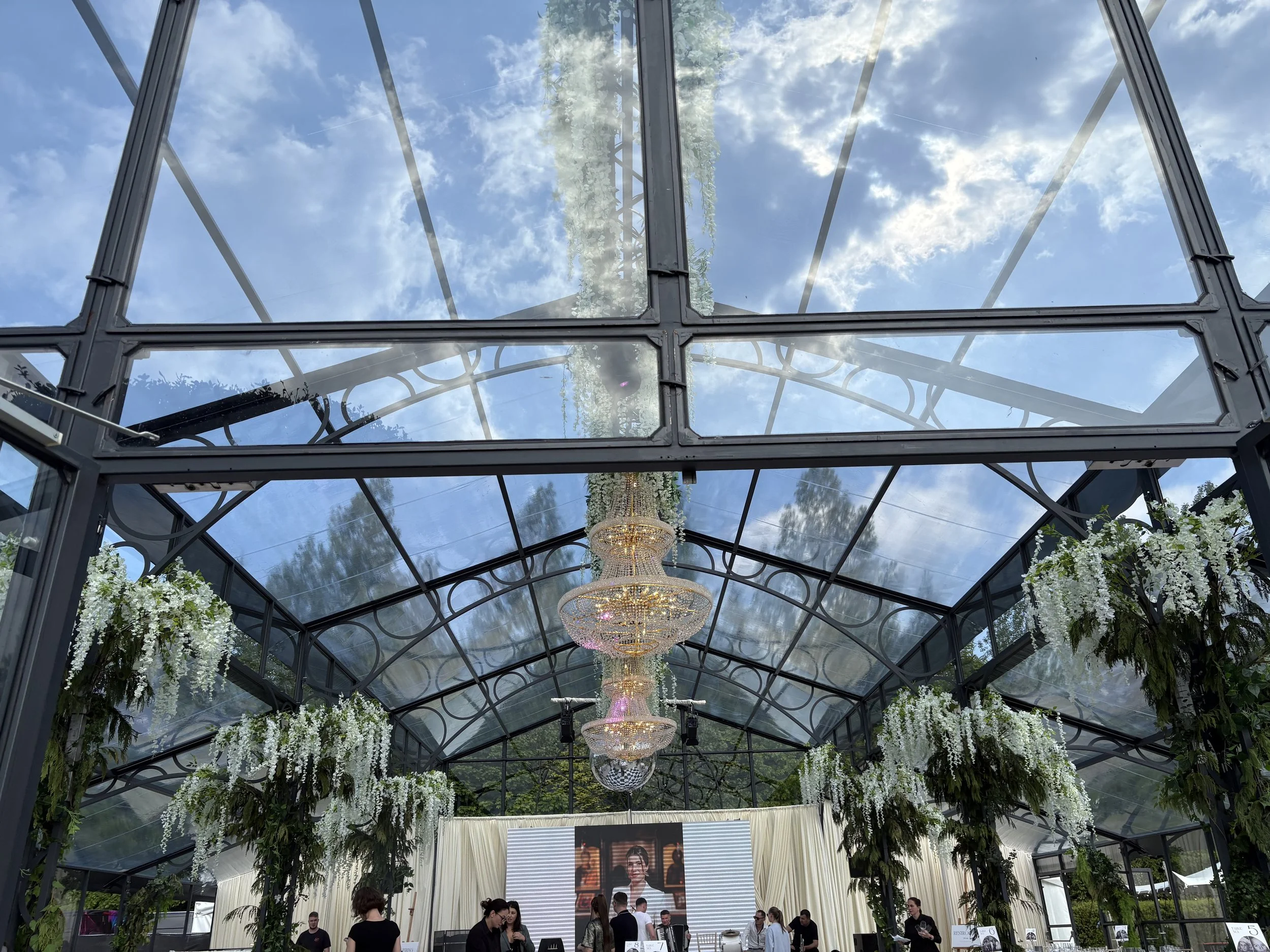 Interior view of a glass greenhouse with a clear sky visible through the roof. The ceiling has chandeliers and is surrounded by hanging white flowers and green foliage. People are gathered inside, with a large screen displaying a person at the front.