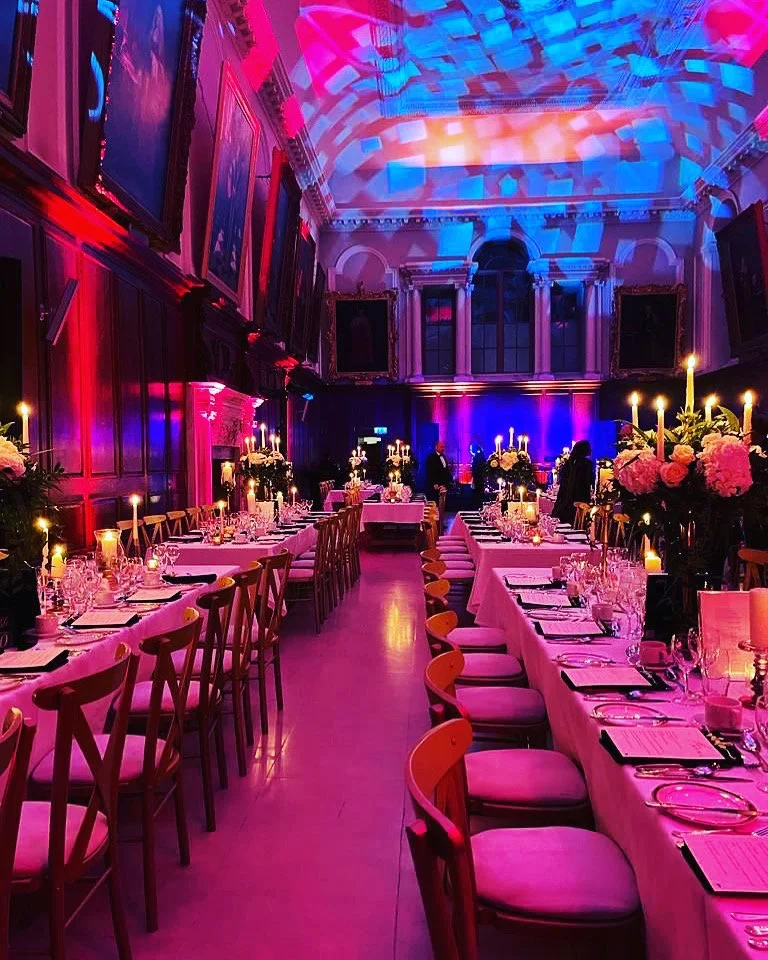 A banquet hall decorated for a fancy event with pink and purple lighting. Long tables with white tablecloths, pink flowers, candle centerpieces, and place settings are set for dining. There are empty chairs around the tables, and a few people are sta