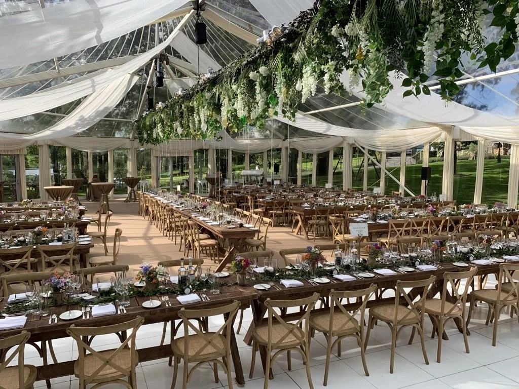 The combination of classic chandeliers, soft florals, and panoramic garden views gives the room a refined yet natural feel, as though the celebration is taking place in an elegant greenhouse in the heart of a landscaped estate.
