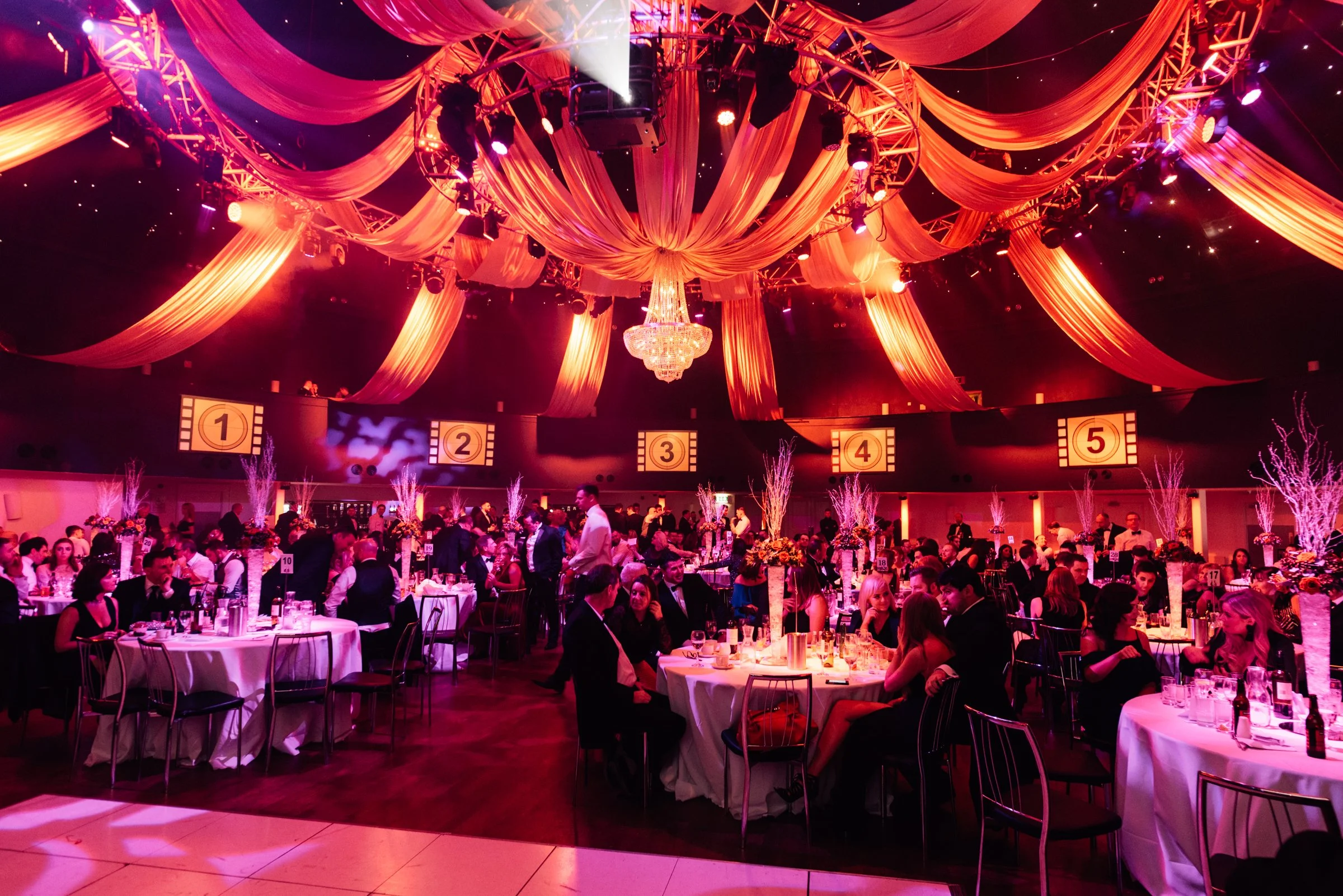 An elegant event hall with pink and purple lighting, draped fabric on the ceiling, a chandelier, and numerous round tables with white tablecloths, floral centerpieces, and guests dressed in formal attire.