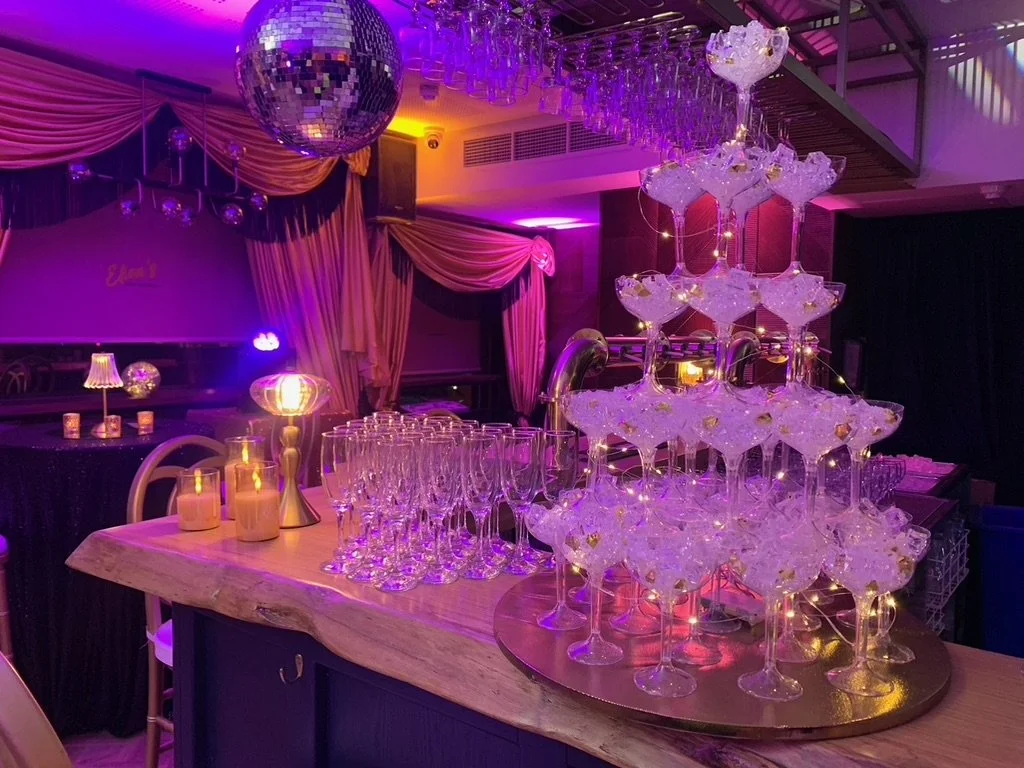 A party venue with a pyramid of champagne glasses and a disco ball, decorated in pink and purple lighting with draped curtains and candles.