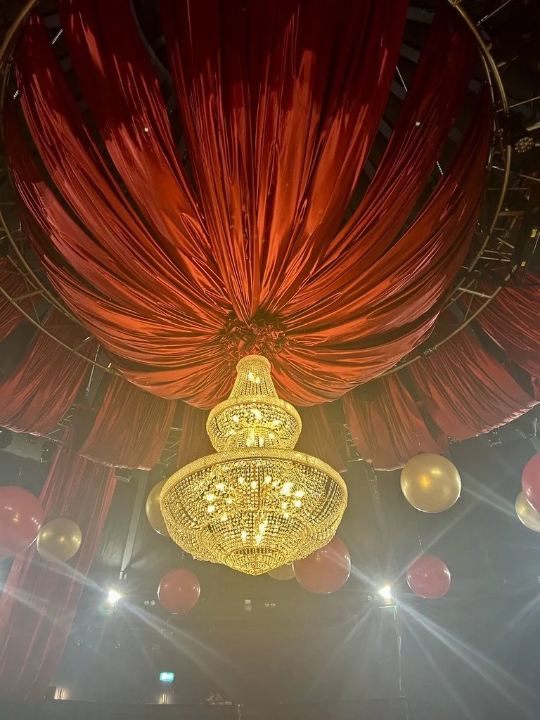 Gold chandelier hanging from a ceiling with red draped fabric and red and gold balloons