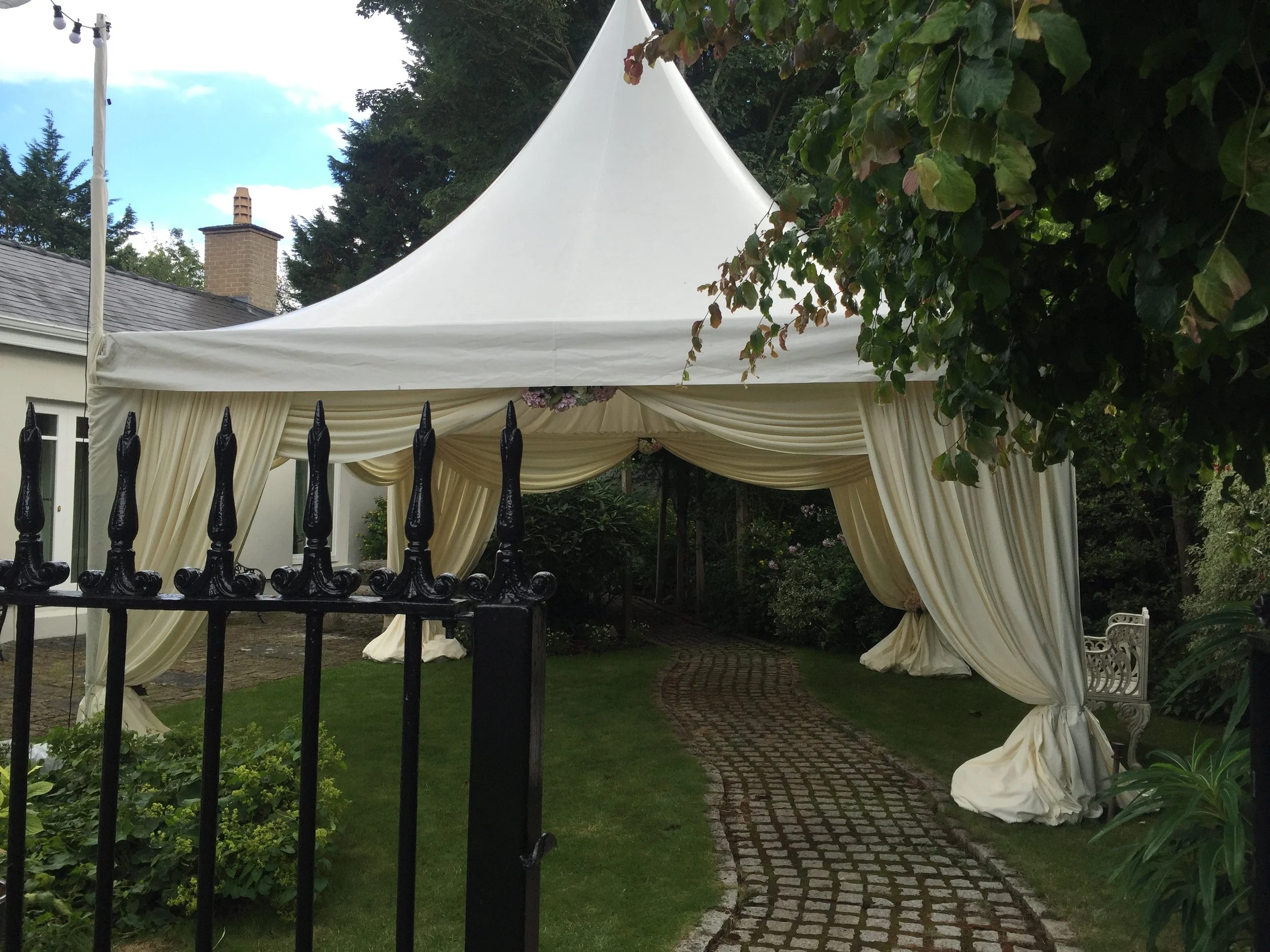 A white event tent with draped sides set up in a garden, surrounded by greenery and trees, with a brick pathway leading underneath.