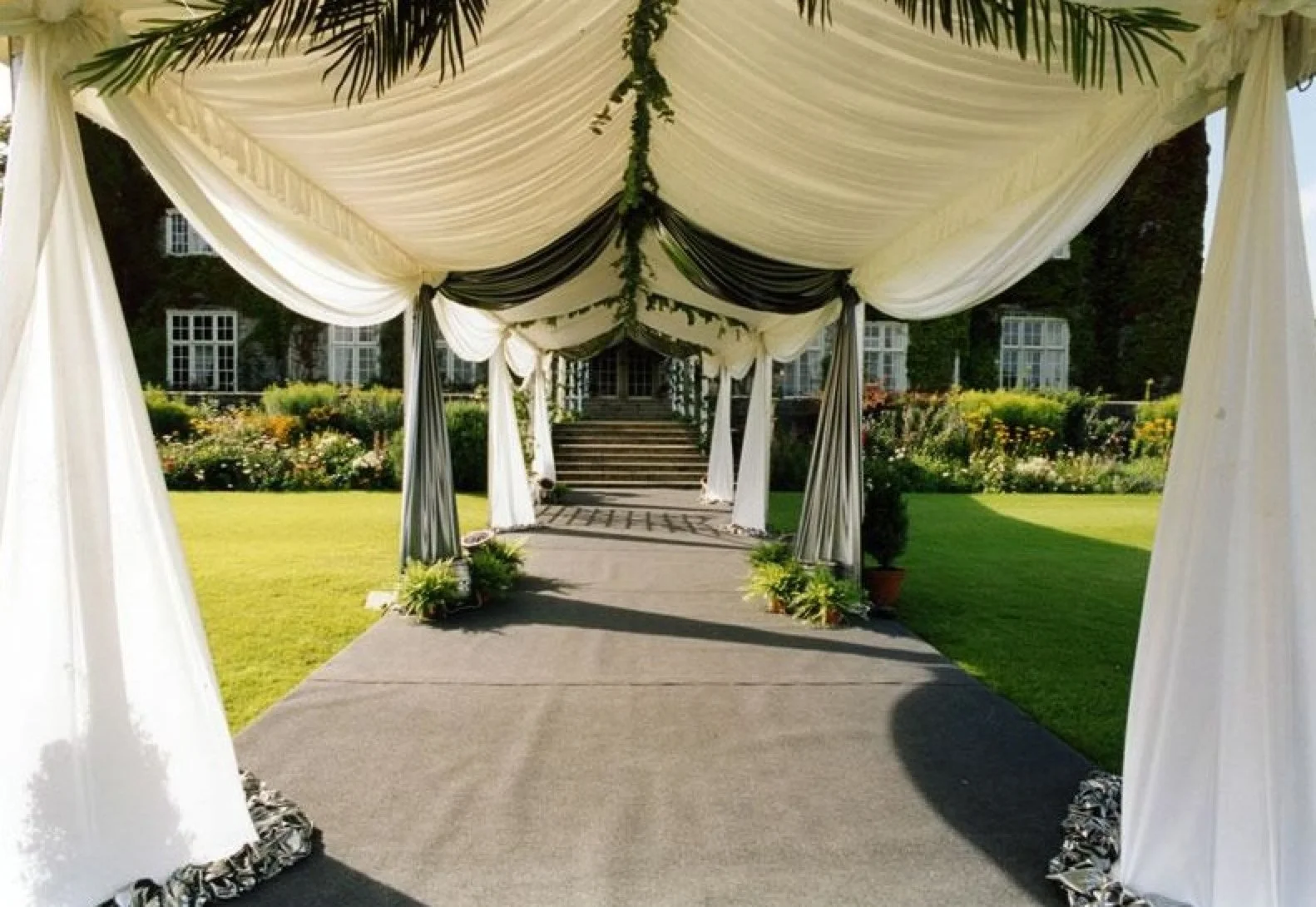 A white wedding tent with black accents, decorated with flowers and greenery, set up on a lawn with a house and garden in the background.