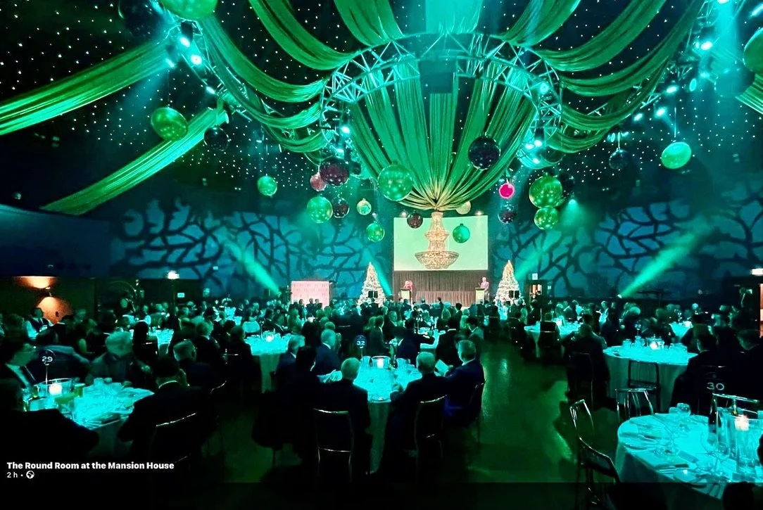 A large banquet hall decorated with green drapes and hanging ornaments, filled with round tables and seated guests, with a stage in the front and dramatic lighting.