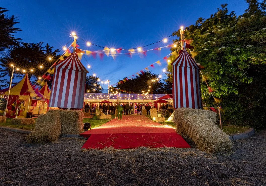Circus-themed outdoor event at dusk with striped tents, string lights, hay bales, and a red carpet leading to a gathering of people.