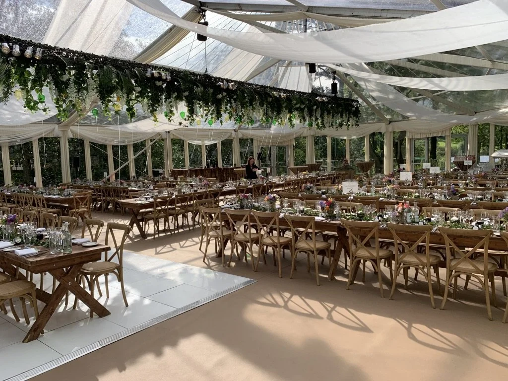 Elegant event space with wooden tables and chairs, floral centerpieces, and decorative drapery in a glass-ceiling tent