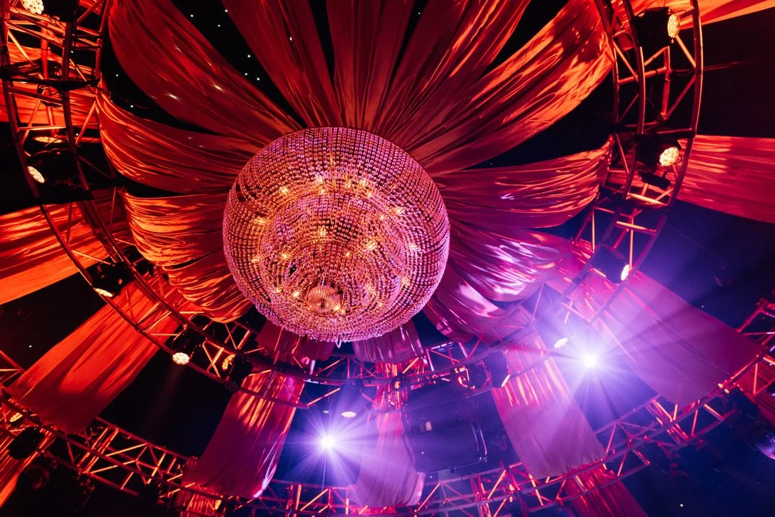 A large, decorative chandelier hanging from the ceiling, surrounded by red drapes and lighting rigging in a dimly lit venue.