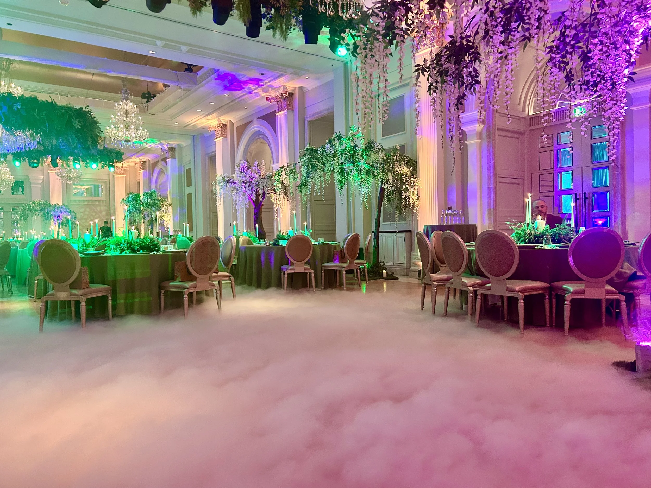 This image captures a ballroom transformed into a dreamy enchanted‑garden setting. Billowing low‑lying fog rolls across the floor like clouds, turning the central space into a floating clearing between the dining tables.