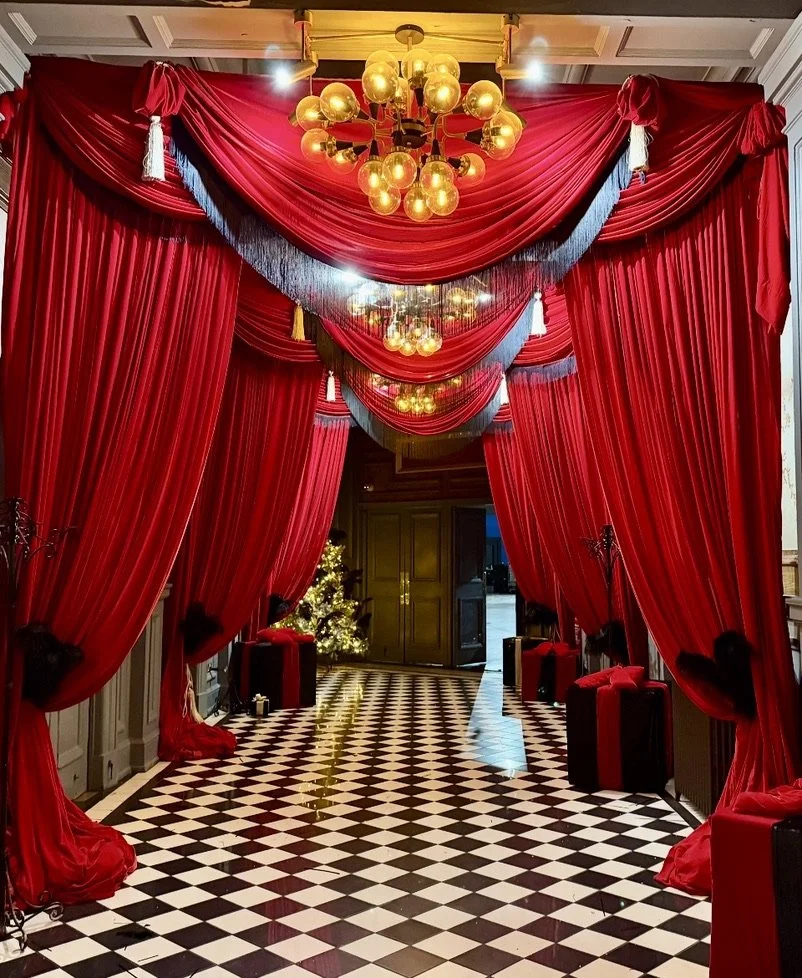 A luxurious hallway decorated for Christmas with red velvet curtains, gold chandeliers, and a decorated Christmas tree at the far end.