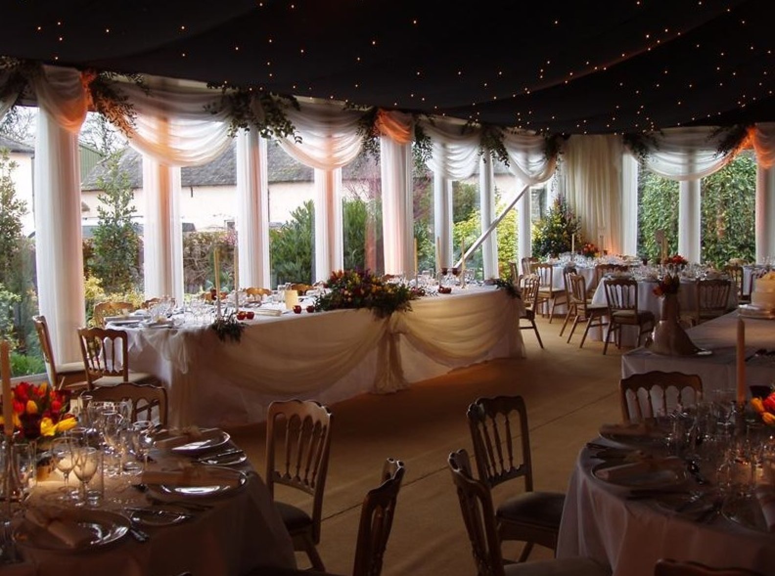 Elegant banquet room decorated with white drapes and string lights, set for a wedding reception or celebration, with tables, chairs, and floral centerpieces.