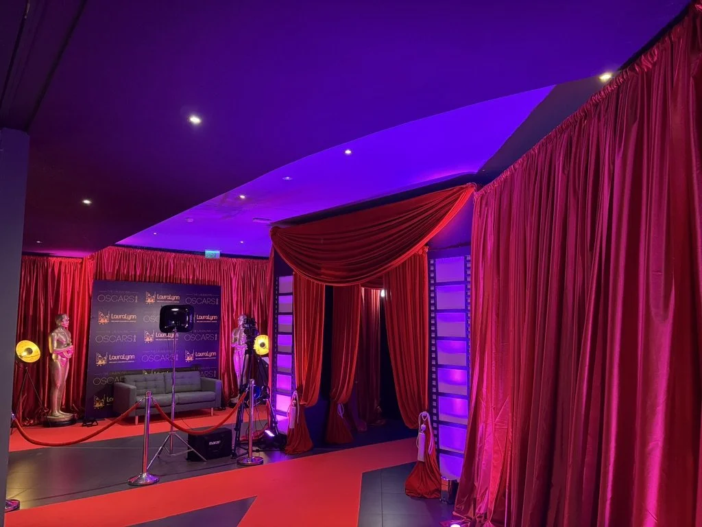 Red velvet curtains and Oscar statues on pedestals at an event venue with a backdrop labeled 'Oscars' and 'Laura Lynn,' with a red carpet and stage setup, and purple lights illuminating the scene.