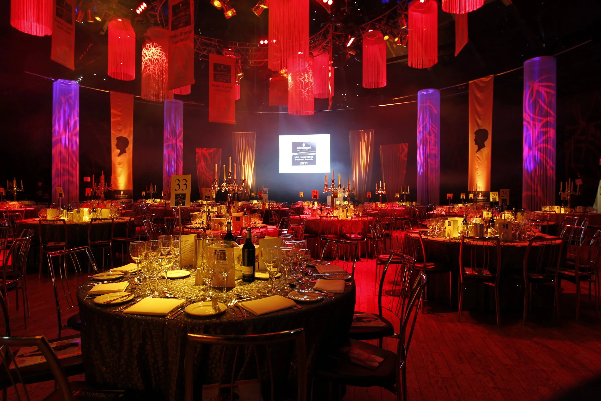 A decorated banquet hall with red and purple lighting, round tables with gold and black tablecloths, wine bottles, candleholders, and place settings, a large screen at the front displaying event details, and hanging banners with silhouettes of a person's profile.