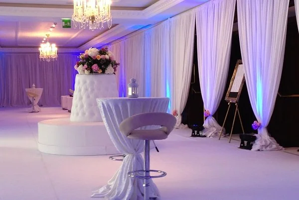 Elegant event space with white drapes, chandeliers, a floral centerpiece on a white chair, and framed artwork.
