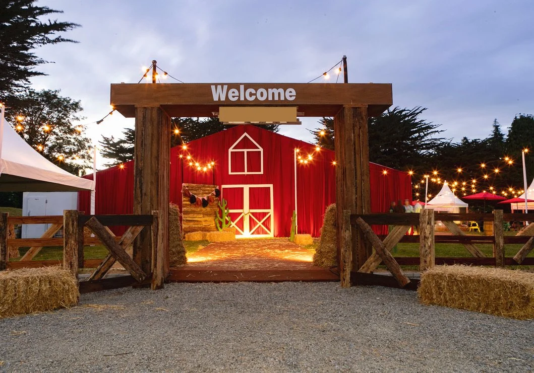 Entrance to a rustic outdoor event space with a wooden gate and 'Welcome' sign, decorated with string lights, hay bales, and a barn backdrop.