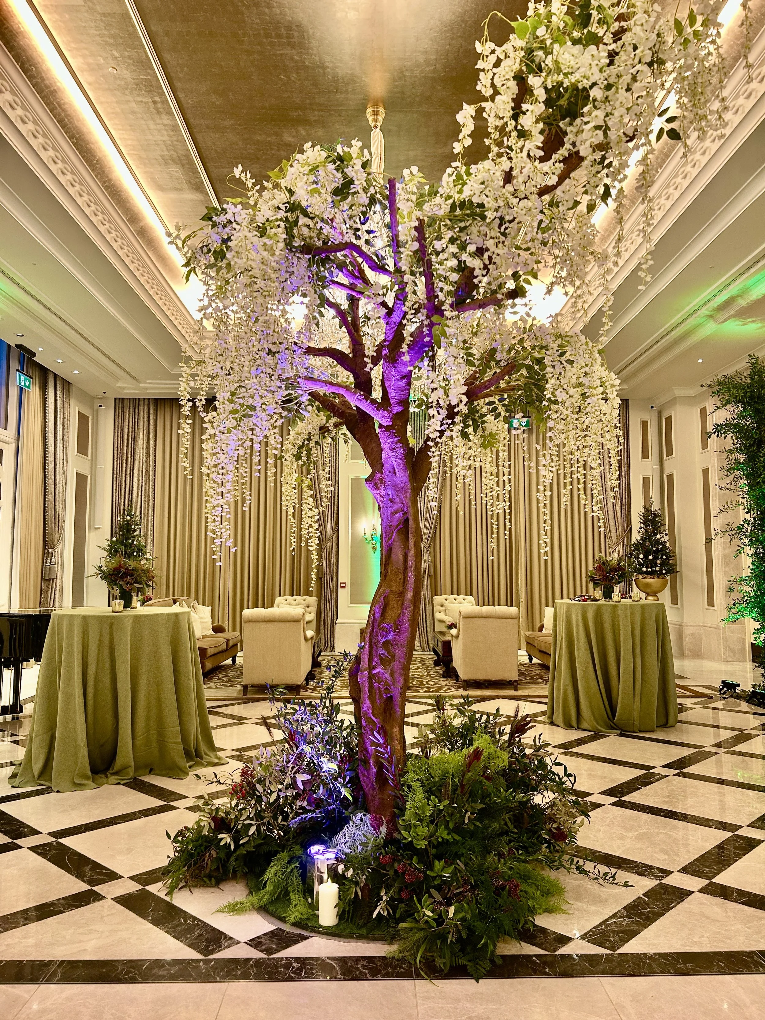 Elegant indoor event space featuring a large artificial tree with white flowers and purple lighting. The room has high ceilings with ornate molding, beige curtains, and a black and white checkered marble floor. There are small round tables with green tablecloths and floral centerpieces, and upholstered armchairs around the room.
