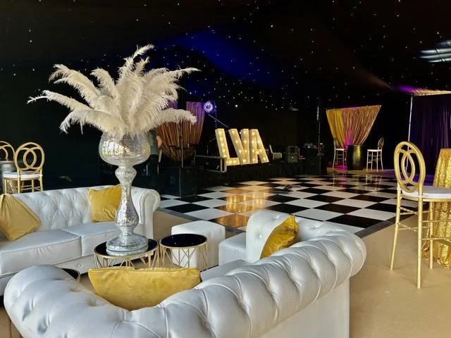 Elegant event space decorated with white sofas with gold pillows, a tall vase with pampas grass, black and white checkered floor, and illuminated marquee letters that spell 'LIVA' in the background.