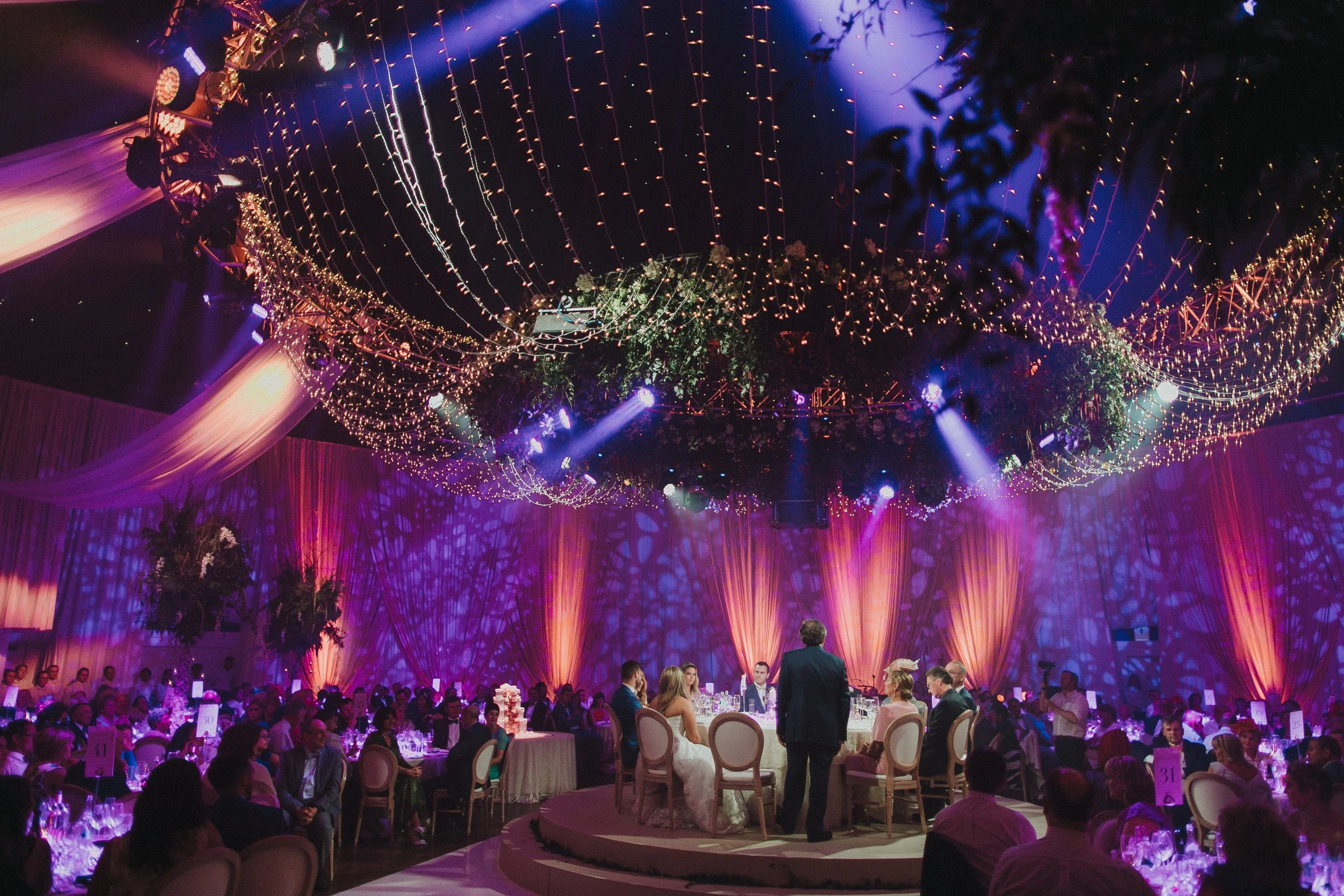 An elegant indoor event with guests seated at decorated tables, a bride and groom at the center, and elaborate lighting and ceiling decorations, including hanging string lights and floral arrangements.