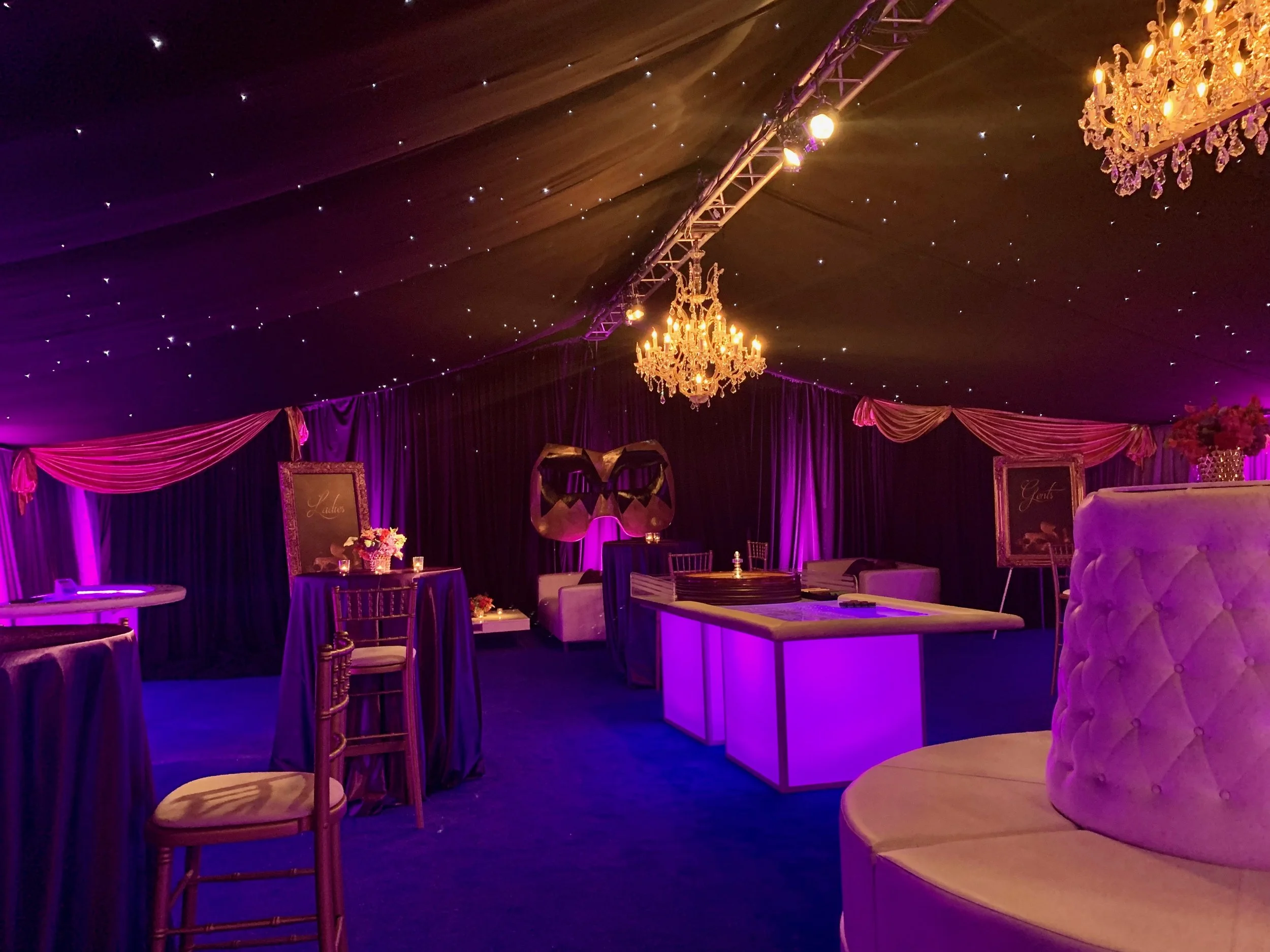 Elegant event space decorated with purple curtains, golden chairs, floral arrangements, and a large chandelier, illuminated with soft purple and pink lighting.