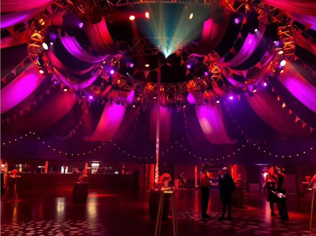 Indoor circus tent with purple, pink, and green lighting, round stage in the center, and a few people standing and conversing inside.