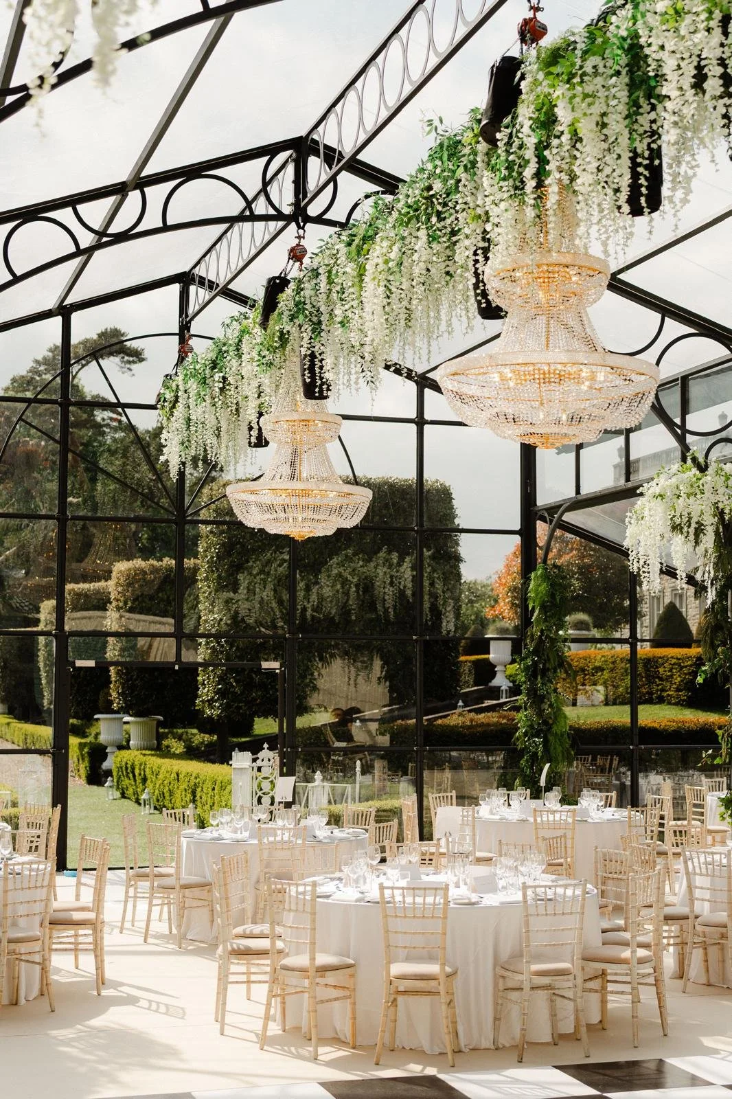 This image depicts an elegant garden‑style reception set inside a bright glass pavilion. Sunlight pours through the transparent walls and ceiling, reflecting manicured hedges, lawns, and urns from the surrounding grounds and making the whole space fe