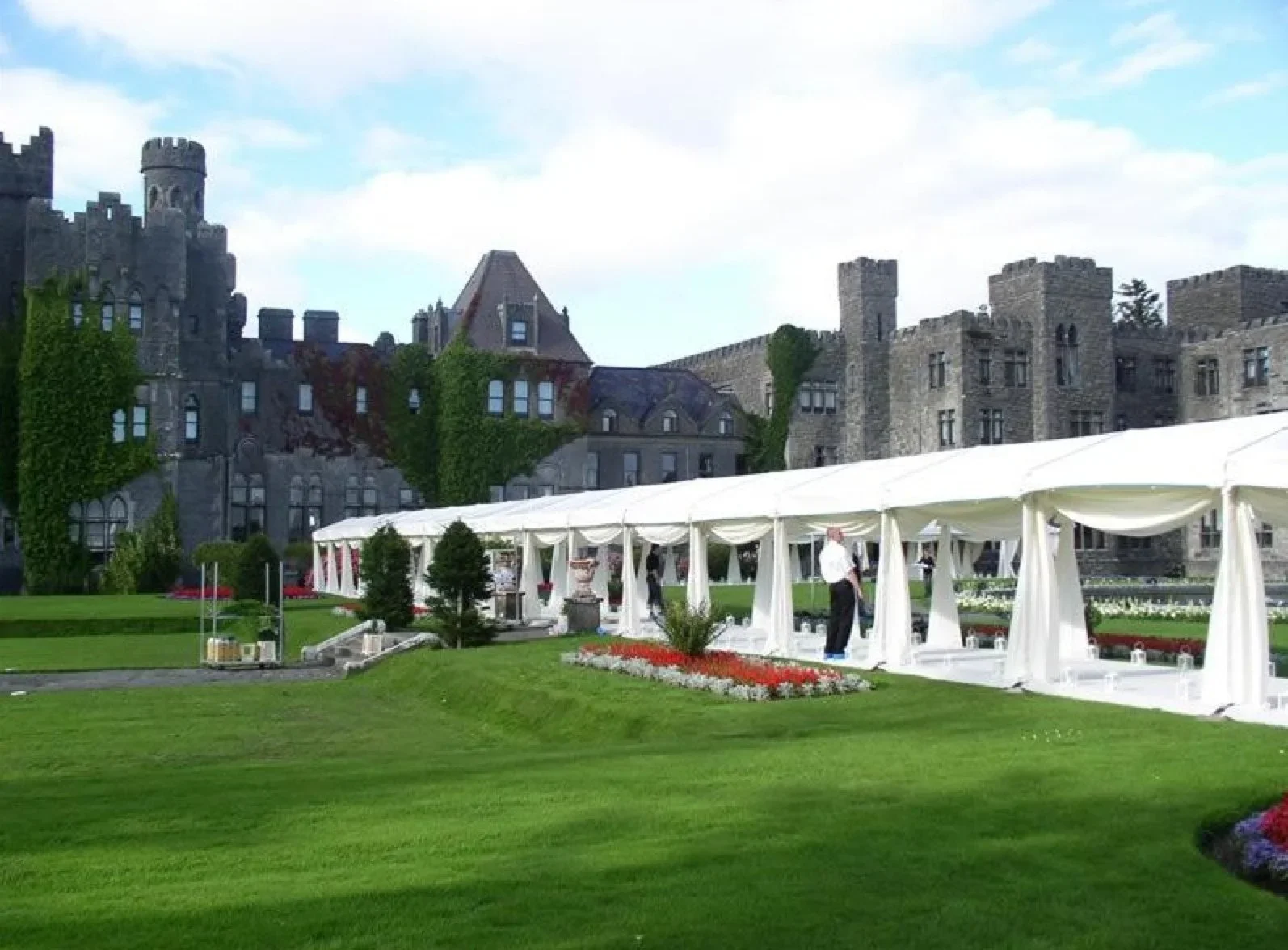 A large outdoor event tent set up on a manicured lawn in front of an old castle-like stone building. The tent is white with draped curtains, and there are people setting up or preparing inside. The background features a historic castle with towers, turrets, and ivy-covered walls.