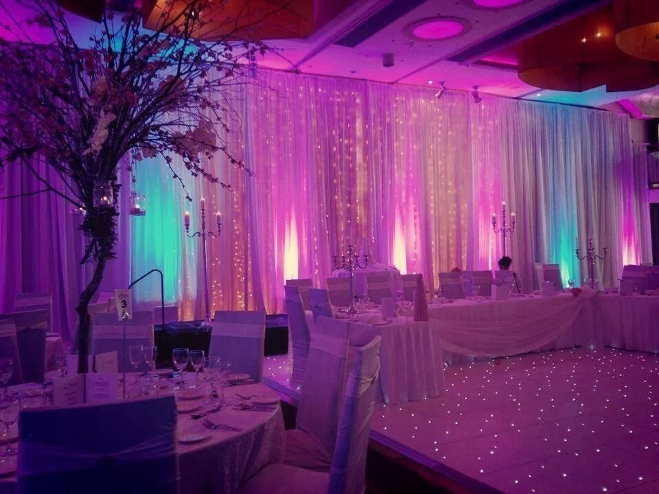 Elegant event space decorated with colorful uplighting, draped fabric, candles, and a floral centerpiece, set for a formal dinner or celebration.