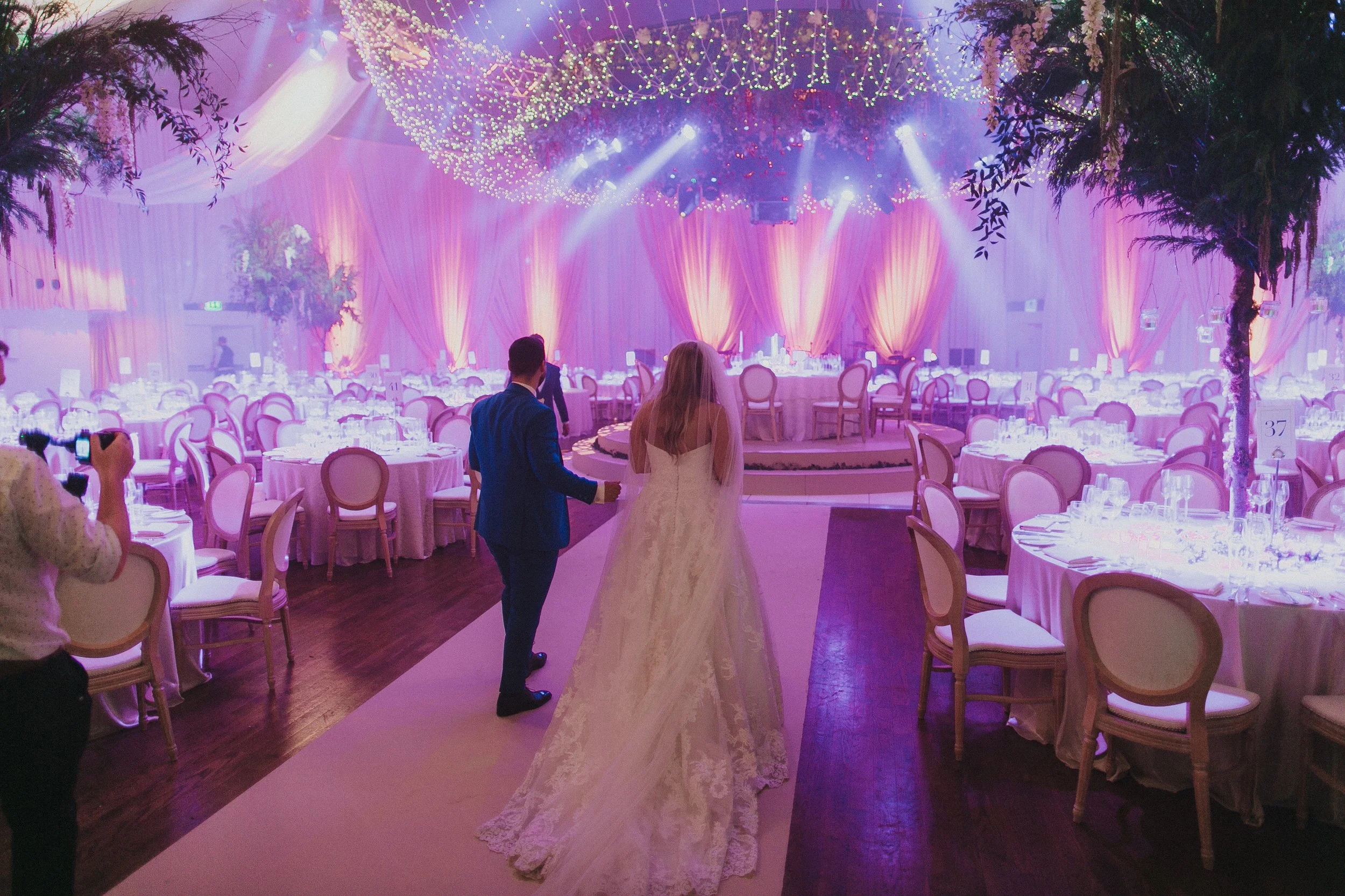 A bride and groom walking down the aisle in a decorated wedding reception hall with pink lighting and round tables with chairs.